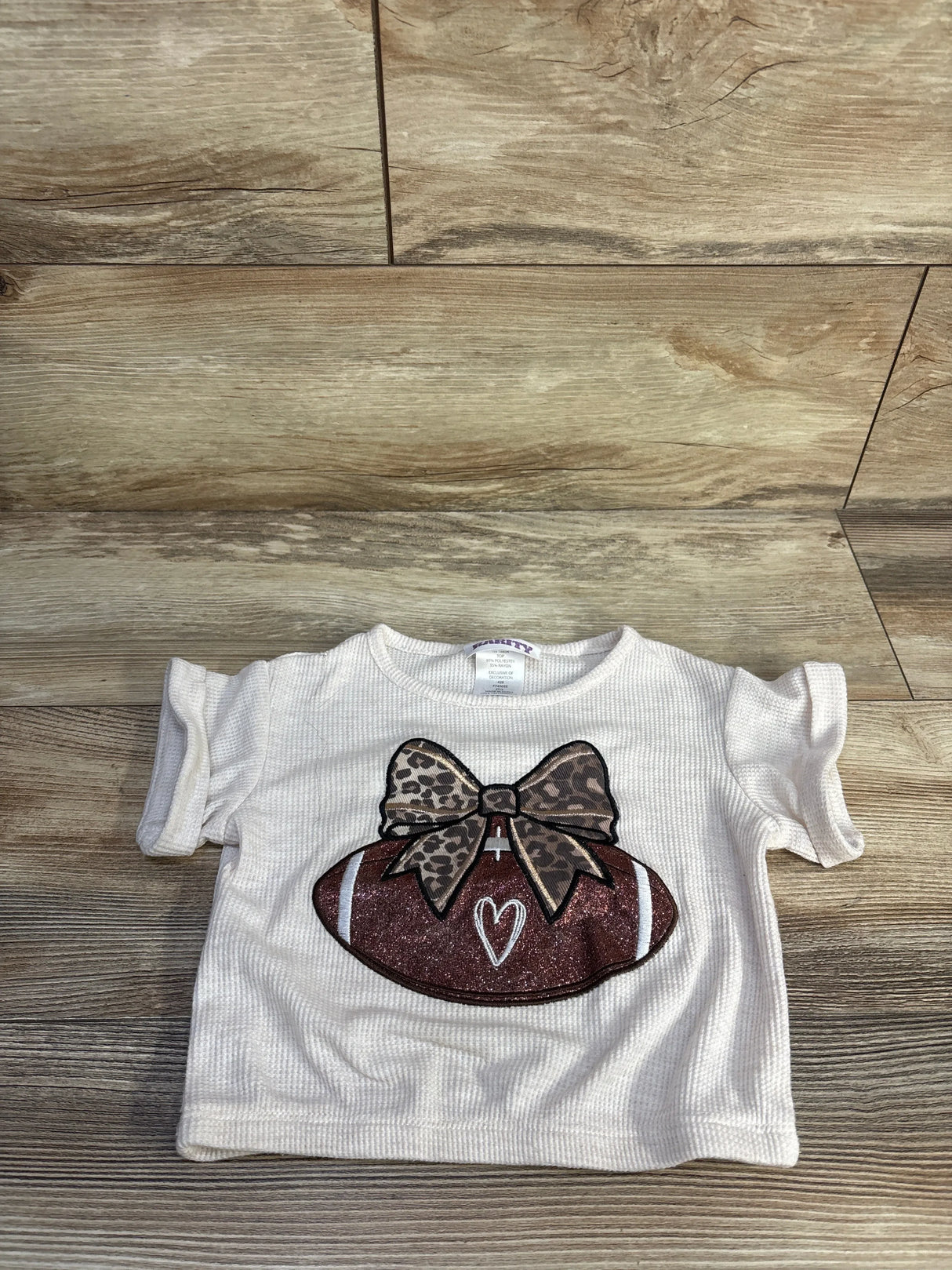 Flat-lay of beige waffle knit toddler football shirt (3T) showcasing glitter football with leopard bow on wood background