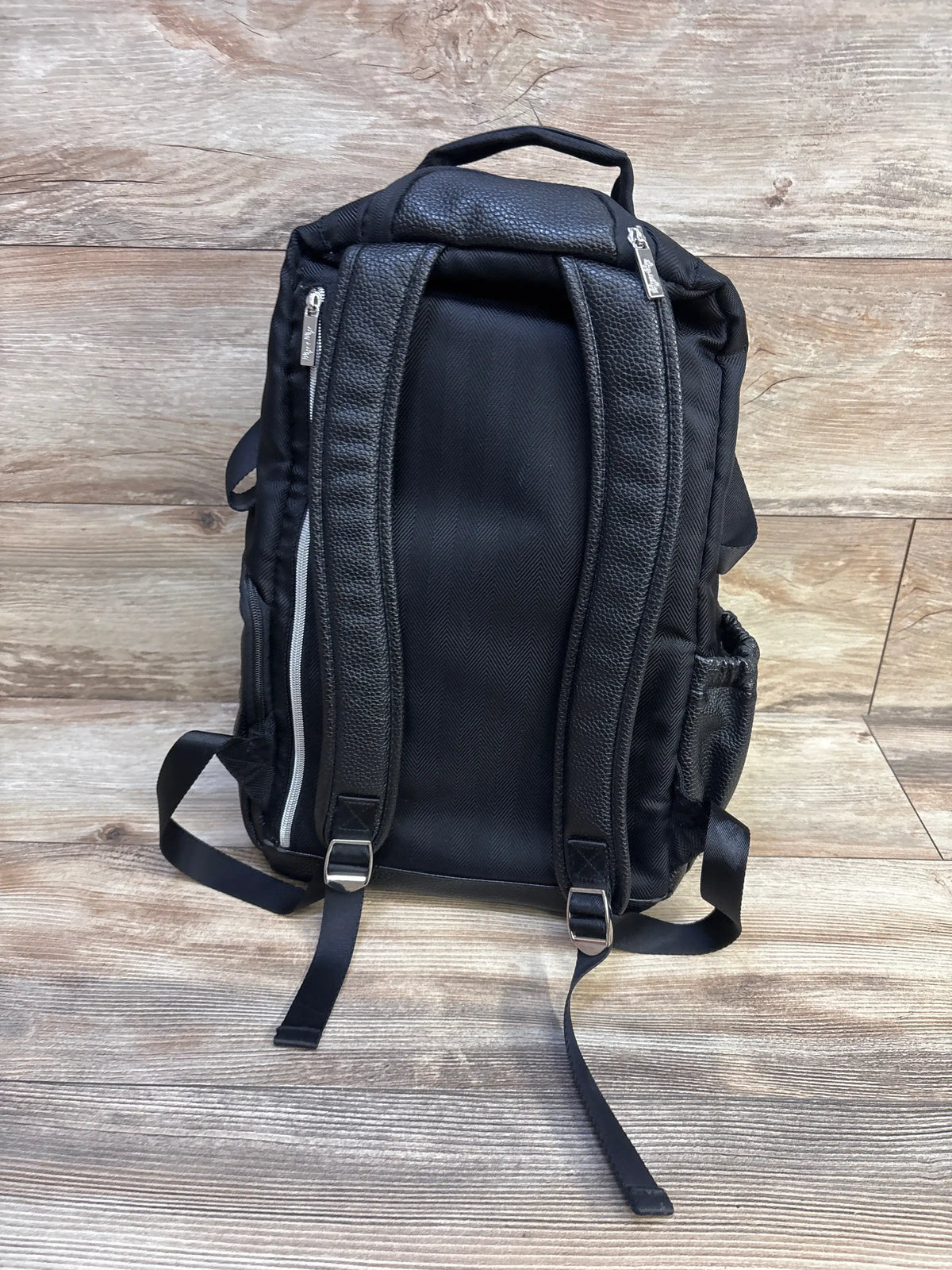 Rear view of Itzy Ritzy Boss diaper bag backpack in black herringbone showcasing shoulder straps and side pocket on a wood floor