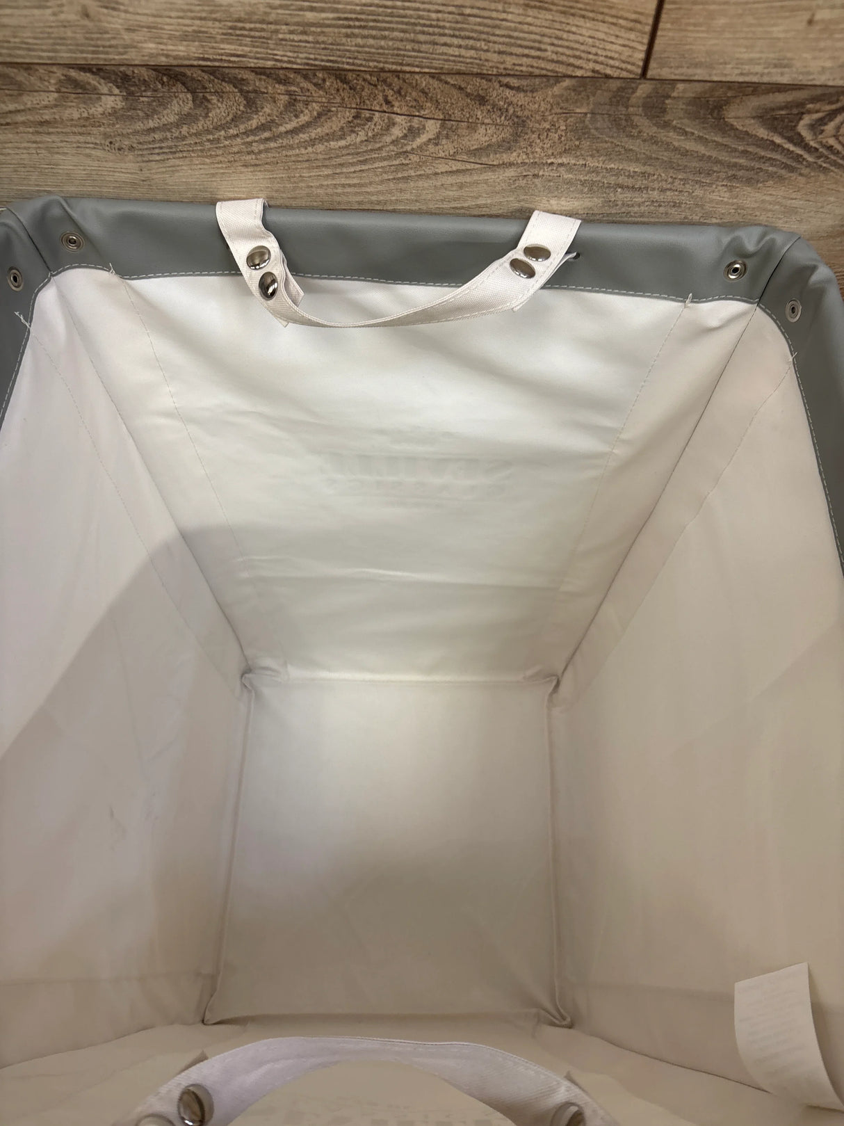 Top-down view of canvas laundry hamper showing white interior, gray rim with metal grommets and snap-on handles on wood floor