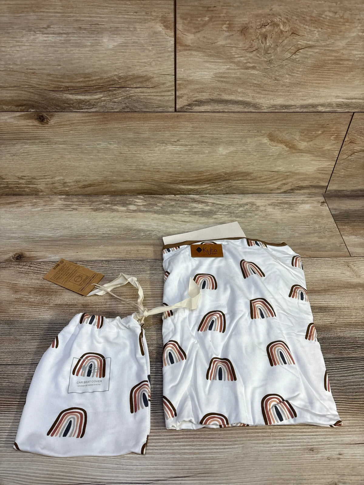 Flat lay of Kyte Baby nursing and car seat cover with rust-brown rainbow print and drawstring bag on wood background
