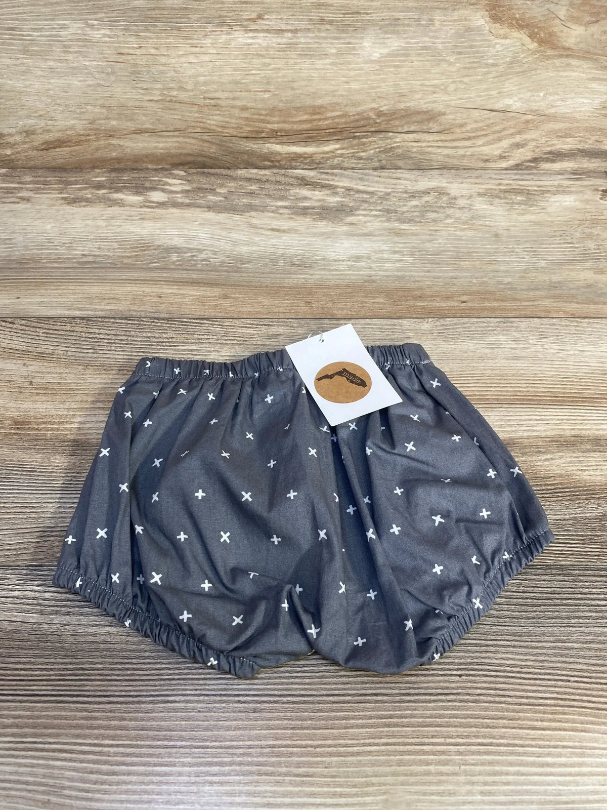 Top-down view of Emerson and Friends grey cotton baby bloomers, size 12 to 18 months, with elastic waist and white cross print on wood background
