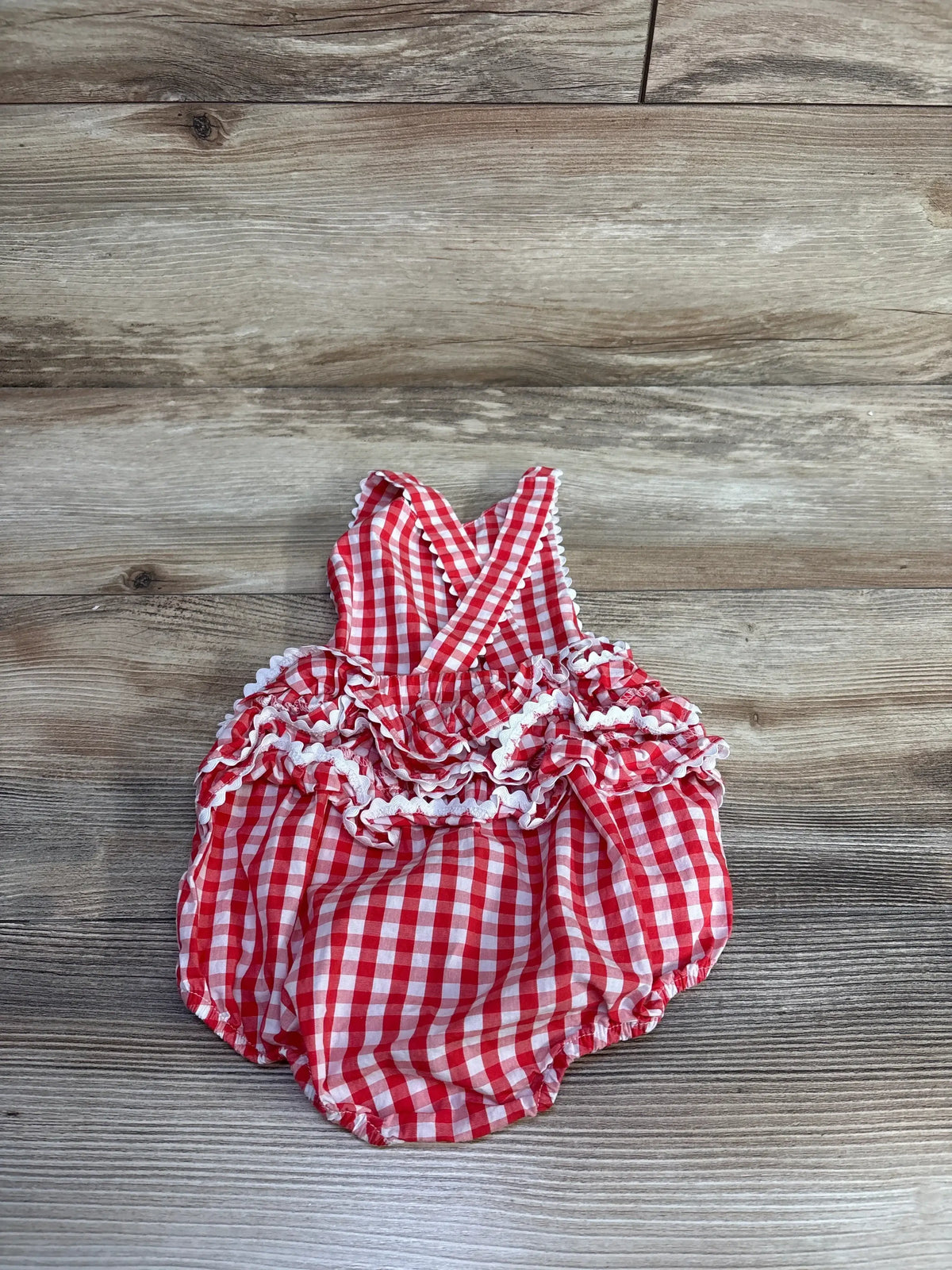 Top down view of red gingham bubble romper showcasing ruffle tiers and crossed shoulder straps on wood plank background