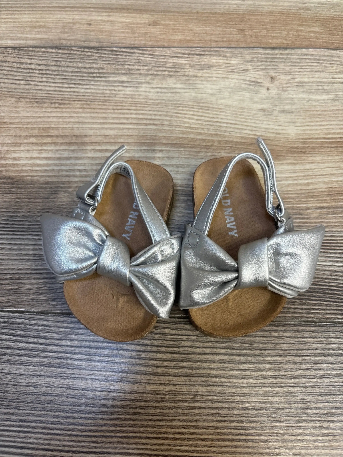 Top view of Old Navy faux-leather tie-bow baby sandals size 1/2C showing silver bows, slingback straps, and brown footbeds on wood