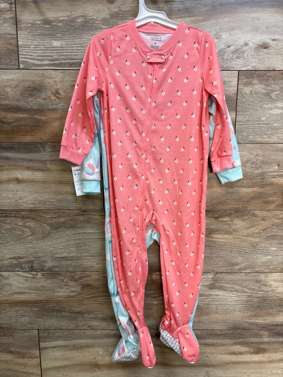 Front view of a two-pack of unicorn-print baby sleepers in pink and blue, with footed legs and zippers on a wooden surface
