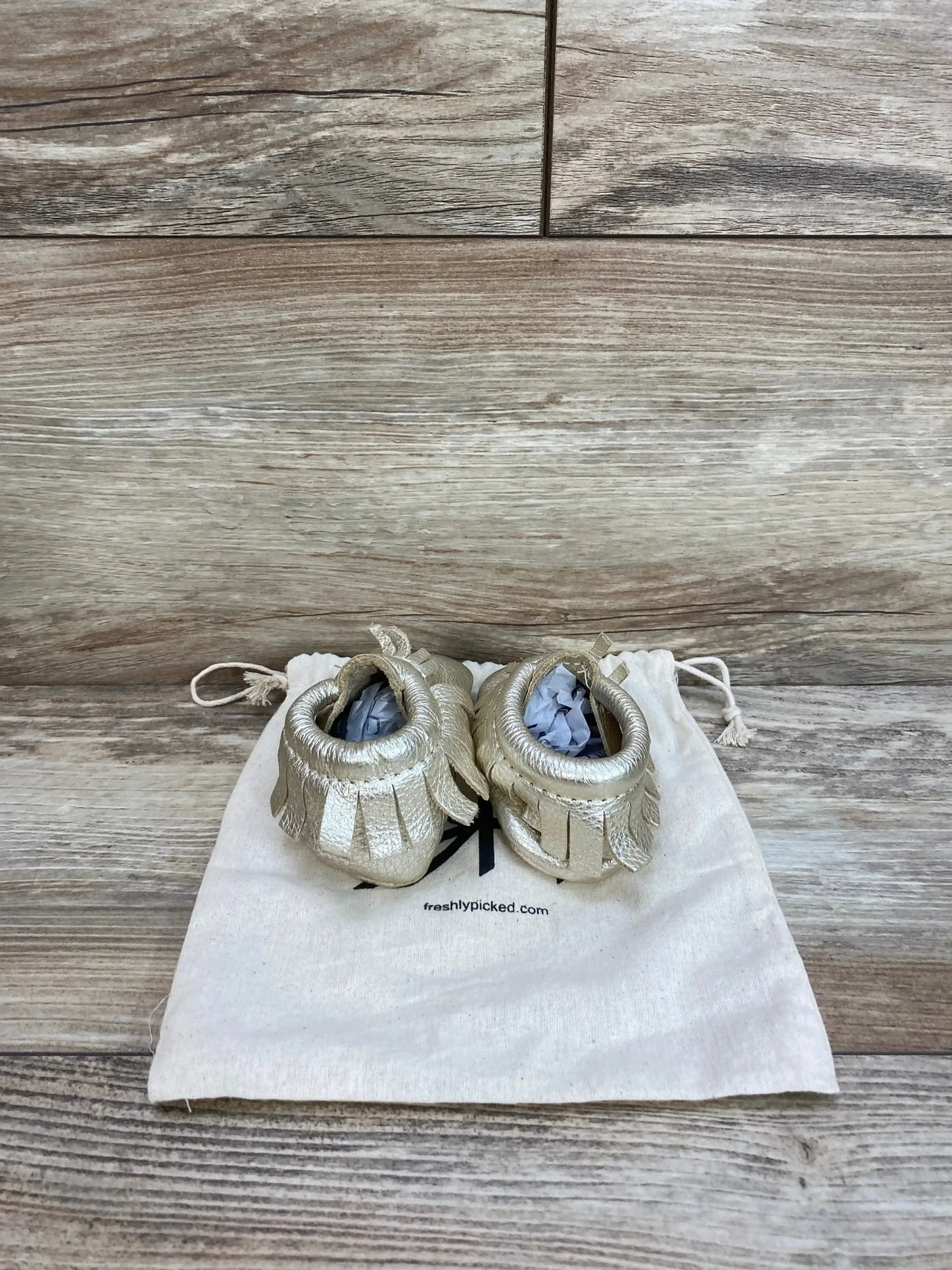 Top view of platinum baby moccasin shoes size 2 showing fringe trim and soft leather on a canvas bag and wood background