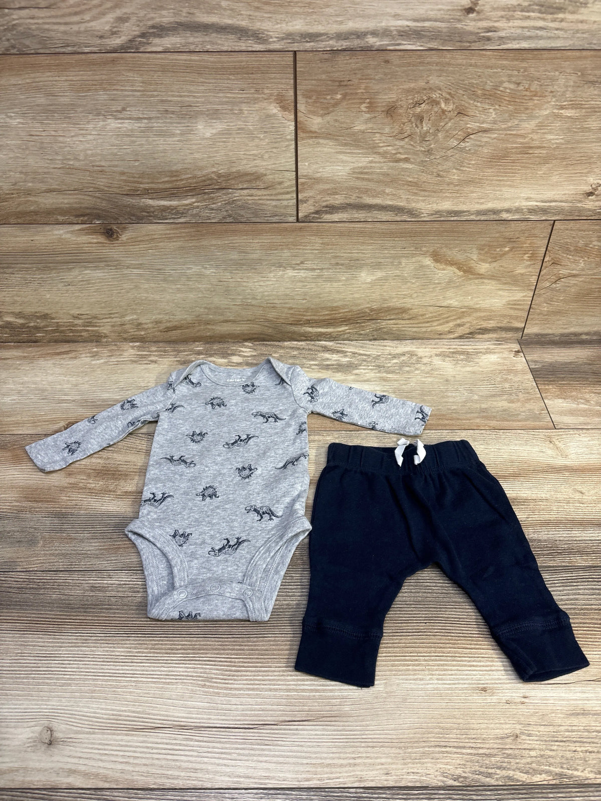 Flat lay of Carter’s two-piece grey long-sleeve bodysuit with dinosaur print and navy elastic-waist pants on wood floor