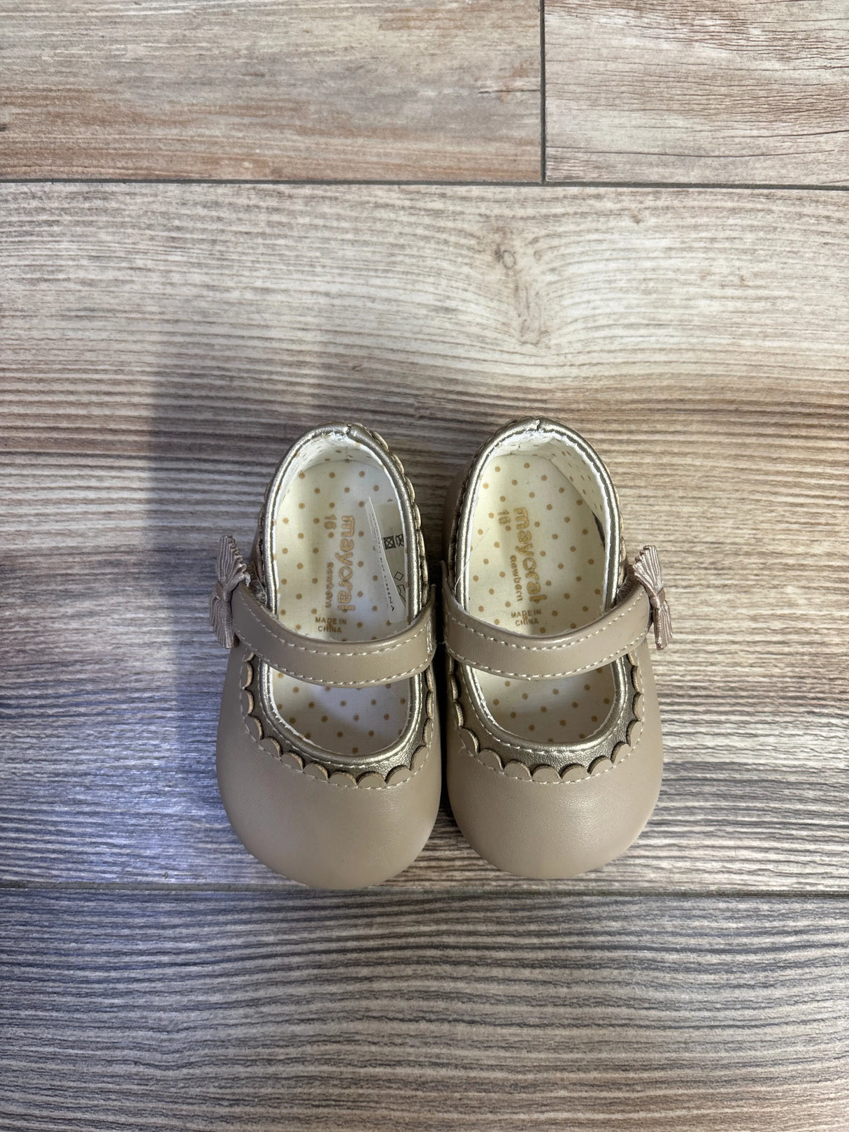 Top view of Mayoral Mary Jane baby shoes in tan showing scalloped metallic trim, strap tassel, and polka-dot insole on light wood floor