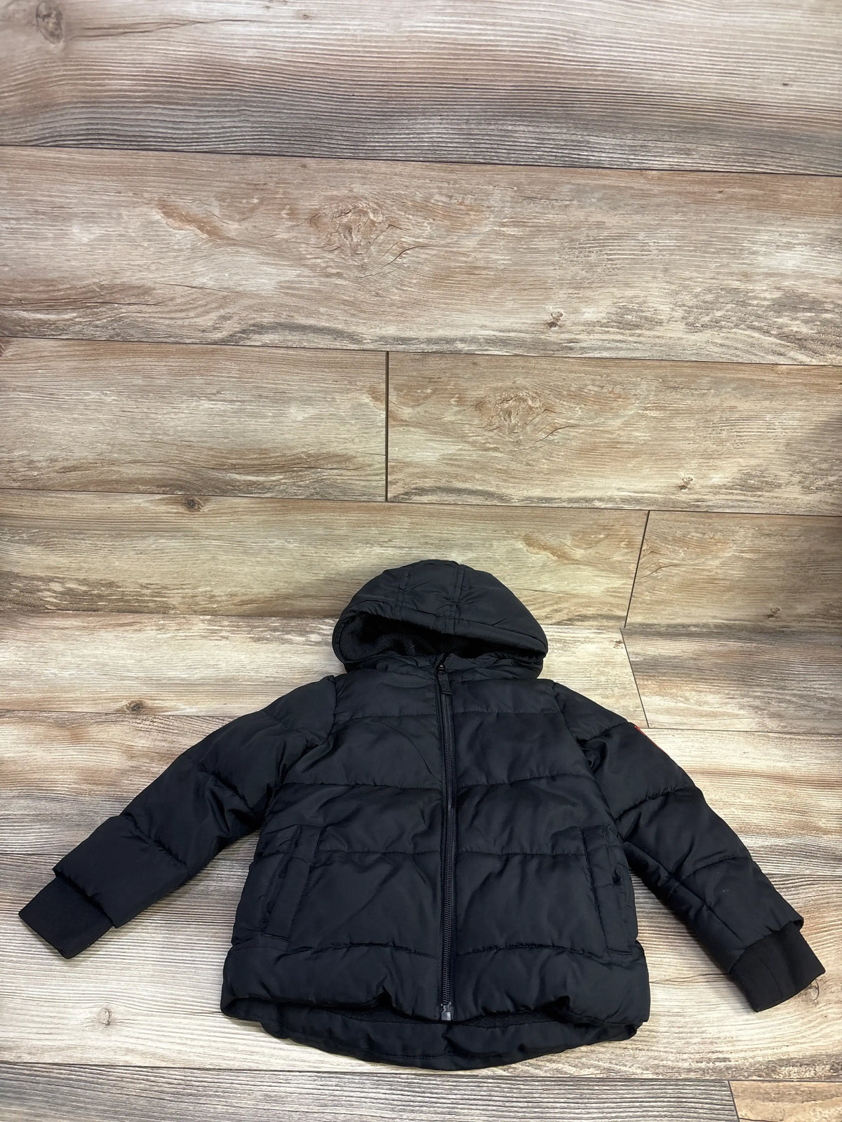 Front view of Okie Dokie puffer jacket in black, size 3T, showcasing hood, quilted panels, zipper, and pockets on a wood floor