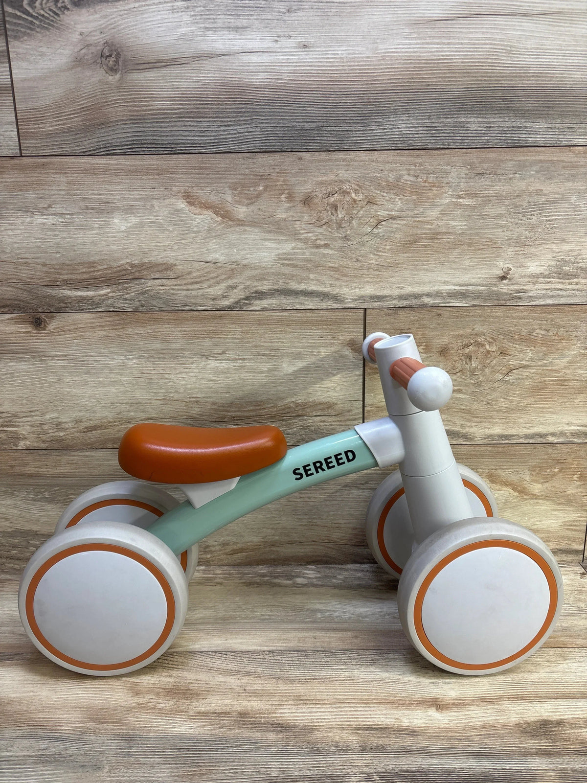 Side view of SEREED balance bike showcasing mint green frame, orange saddle and orange-trimmed white wheels on a wood plank background