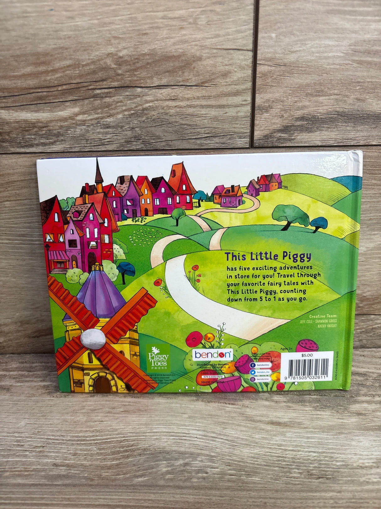 Back of This Little Piggy board book showing colorful village and windmill artwork with barcode and logos on wood floor