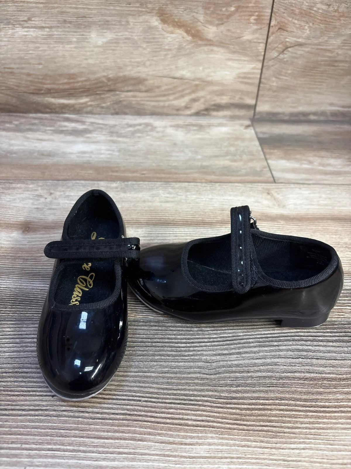 Front and side views of black Dance Class tap shoes, size 7.5C, showcasing glossy finish and secure straps on a wood-grain floor