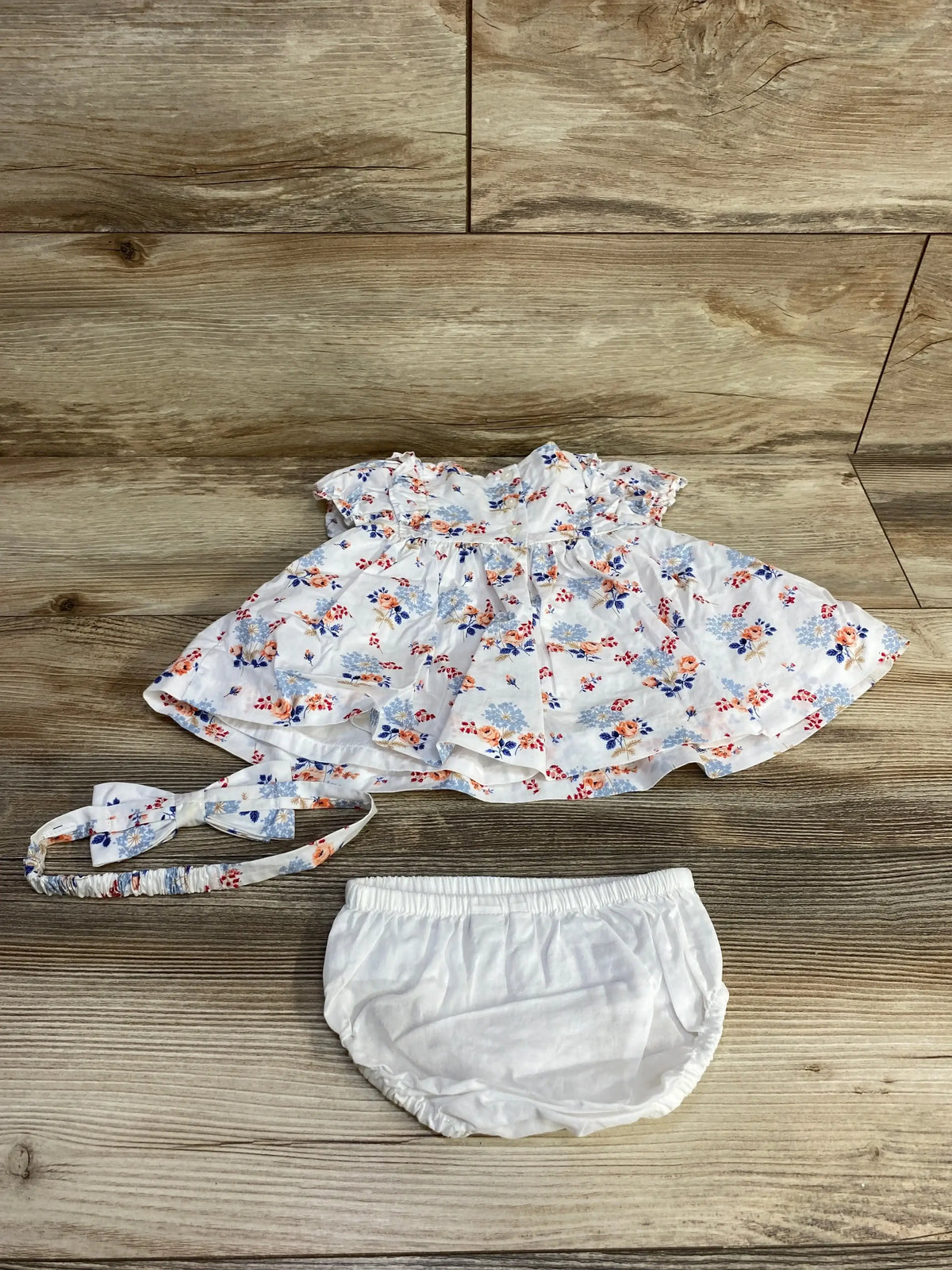 Flat lay of Janie and Jack 3pc Floral Pintuck Dress Set White sz 0-3m showcasing white floral pintuck dress with matching headband and bloomers on wood plank background
