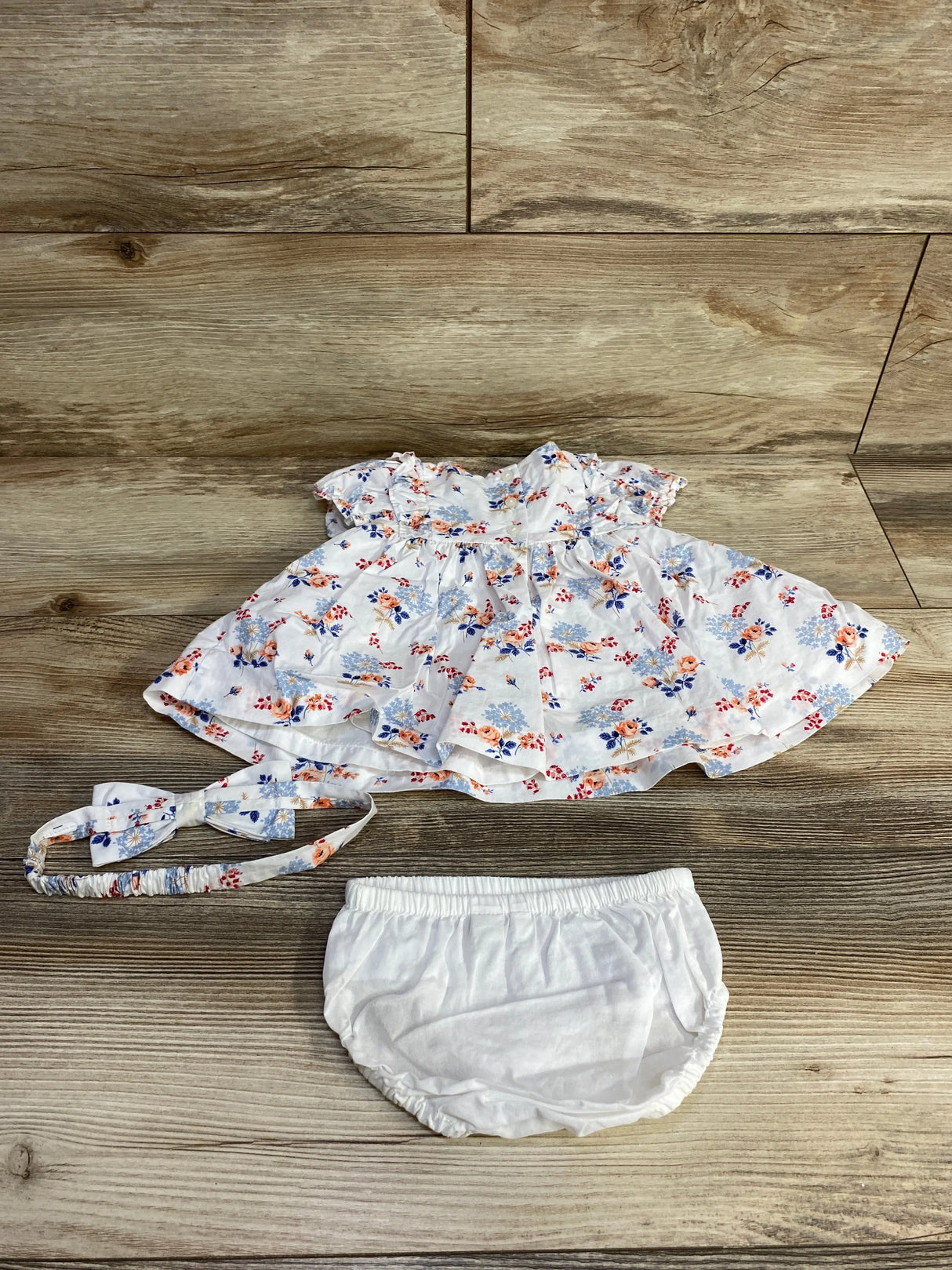 Flat lay of newborn white floral pintuck dress set with matching headband and white bloomers on wood background