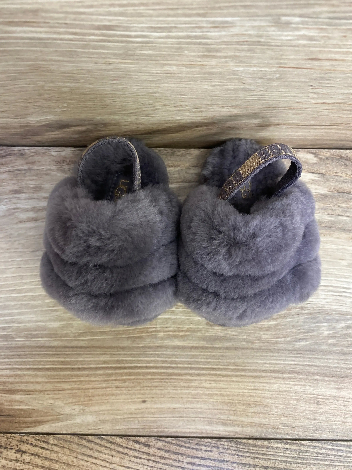 Top view of pair of UGG toddler Fluff Yeah slides in grey showcasing plush sheepskin upper and elastic heel straps on wood floor background