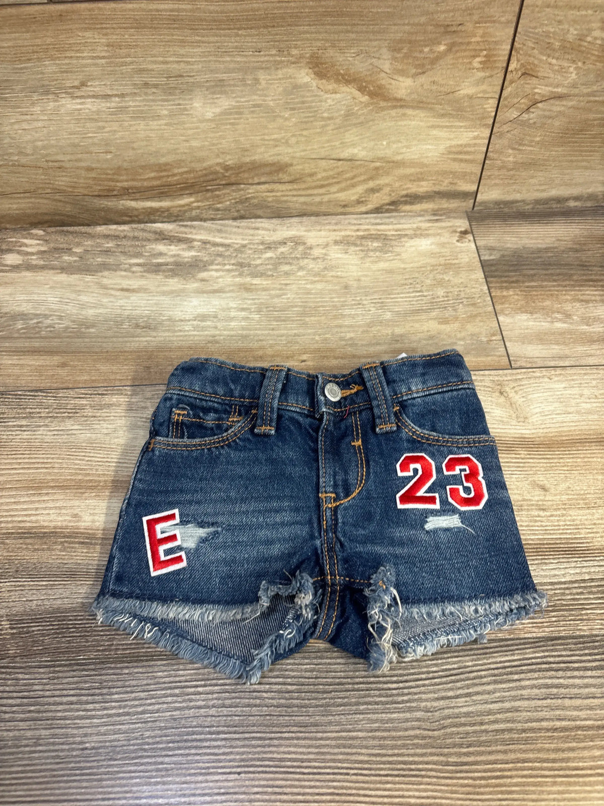 Front view of Old Navy baby denim shorts showcasing frayed hem and red 23 and E patches with wood floor background