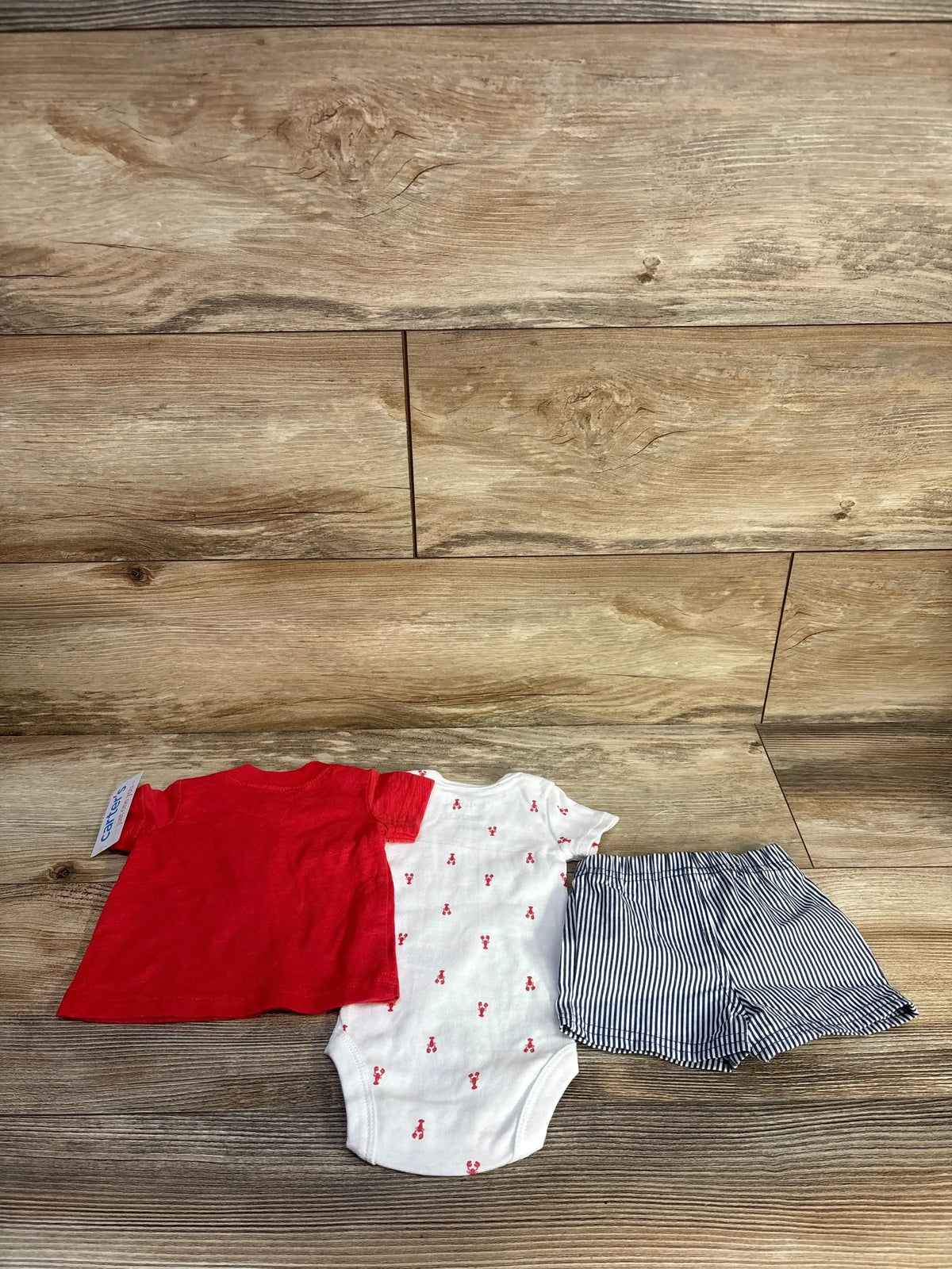 Flat lay of newborn 3-piece set: red henley shirt, white onesie with red lobster print, navy striped shorts on wood