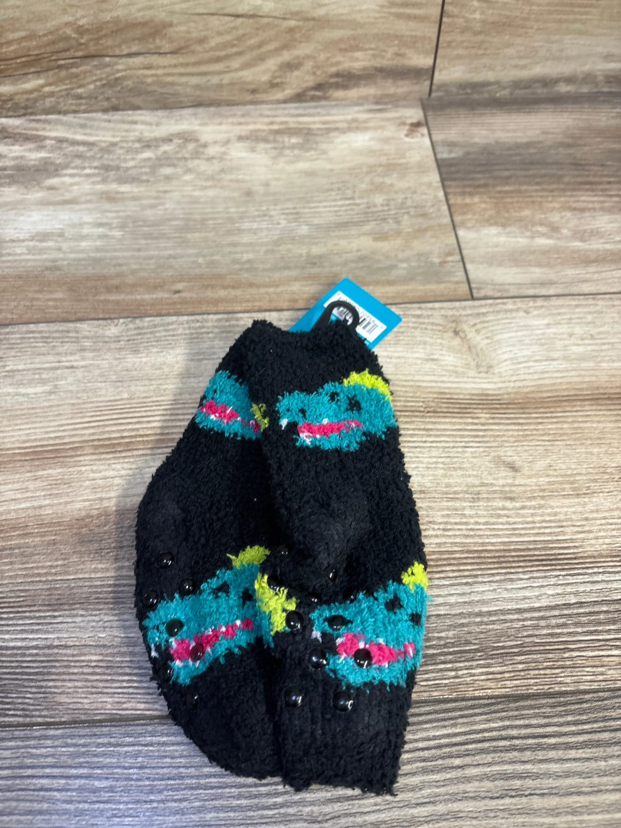 Top-down view of black kids fuzzy socks (West Loop, size 4-8.5C) showing gripper dots and teal dinosaur pattern on wood floor
