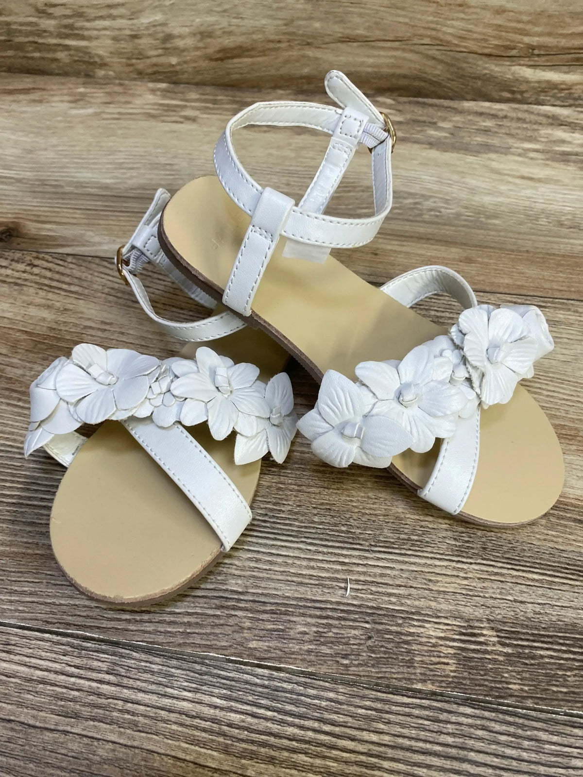 Top down view of white Janie and Jack flower sandals size 12C showing floral straps and ankle buckle on wood background
