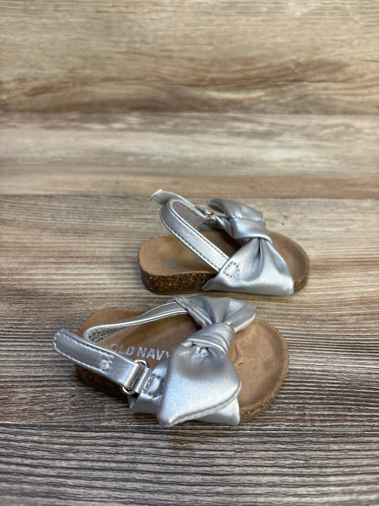 Metallic silver toddler sandals with bow detail and cushioned soles on wood background