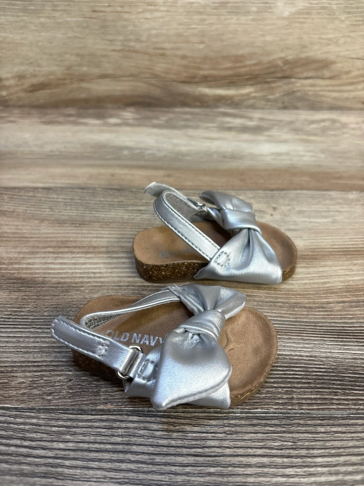 Angled view of Old Navy baby sandals showing metallic silver faux-leather tie bows, slingback strap, and cork footbed on wood background