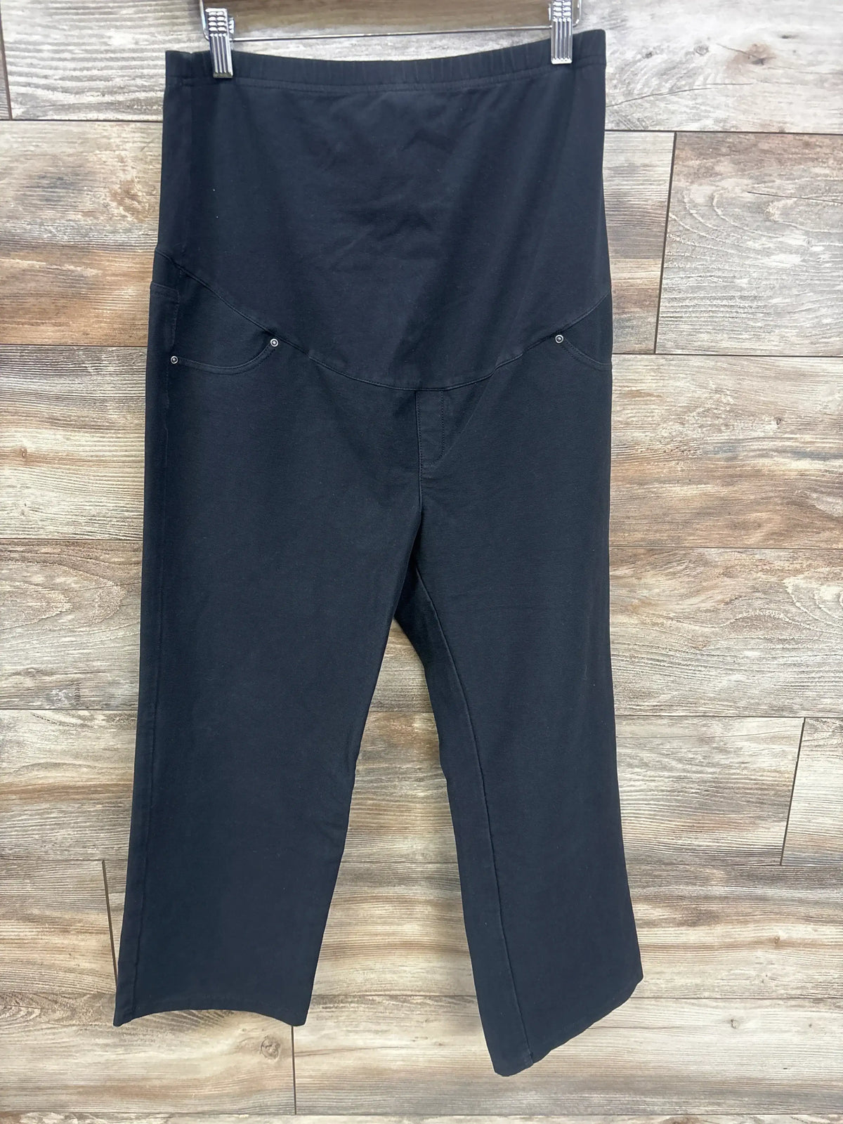 Black maternity pants with over-the-belly panel and side pockets, hanging on hanger against wood floor.