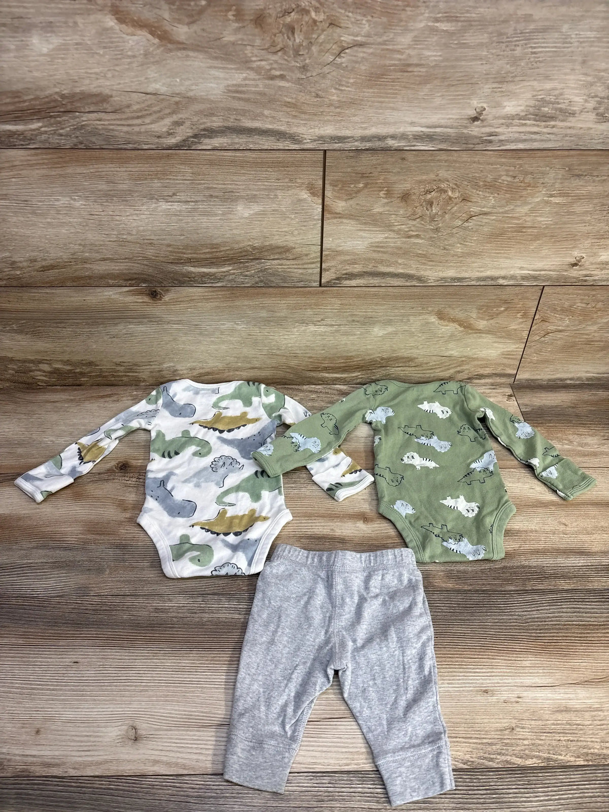 Top view of Carter’s 3pc Striped Bodysuit Set in green, with two dinosaur-print bodysuits and gray pants on a wood floor