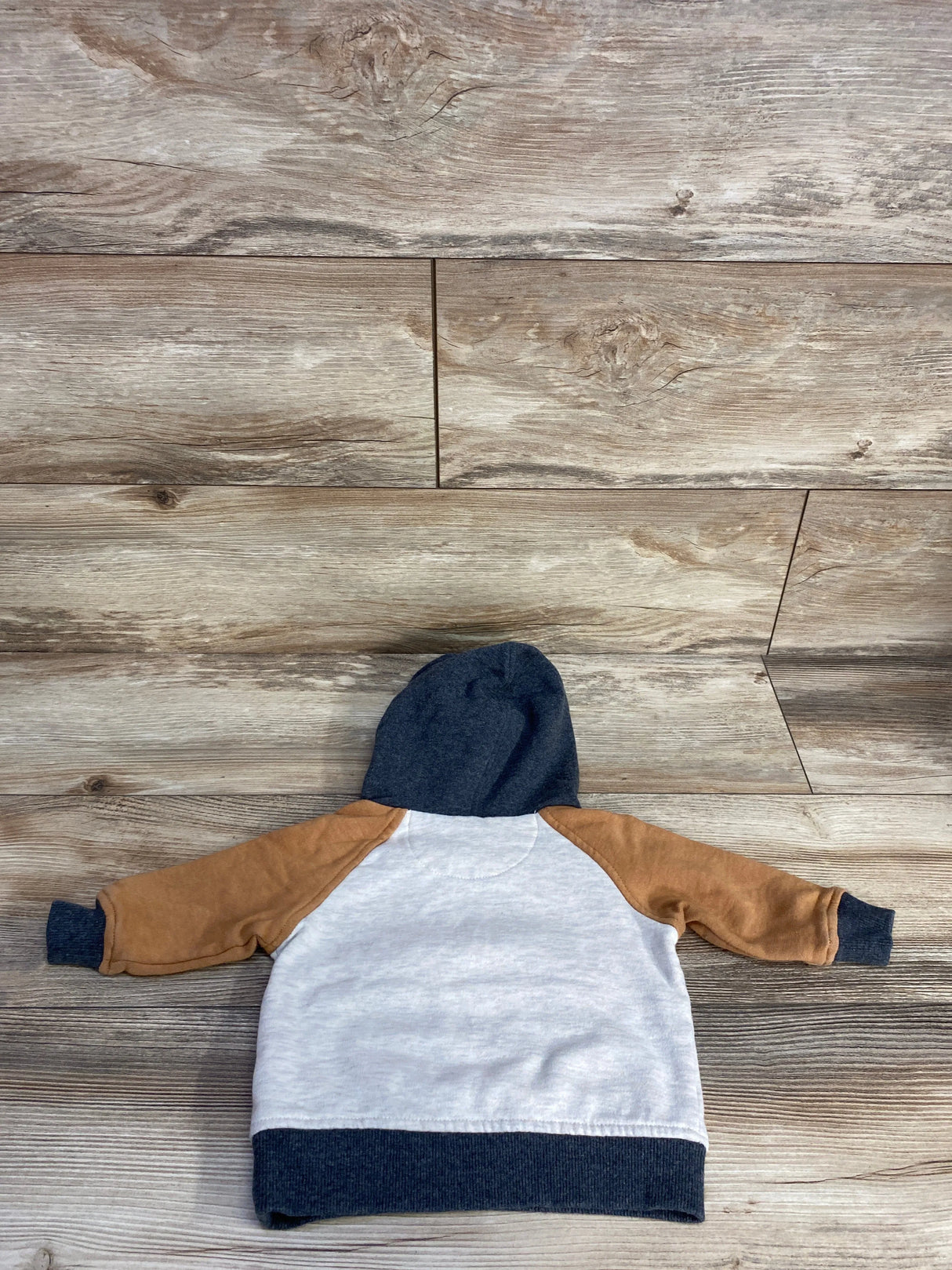 Back view of a kids' hoodie with tan sleeves, white body, and navy blue hood and cuffs on wooden floor