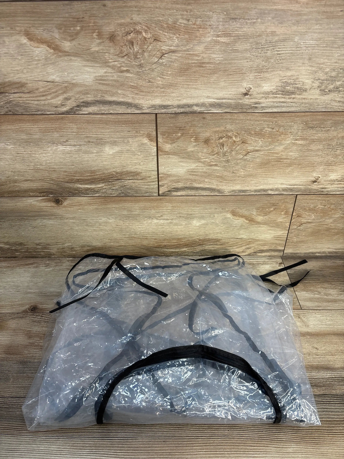 Top-down view of folded transparent stroller and car seat rain cover showcasing black trim and tie straps with wood floor background