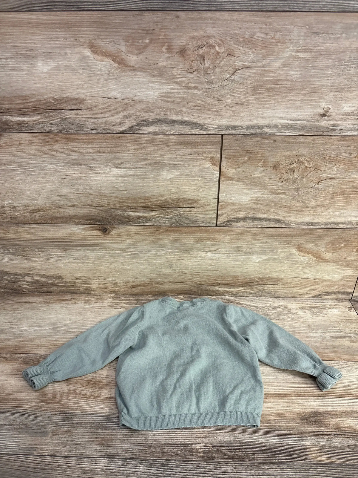 Back view of light green baby button-up cardigan showing knit texture, ribbed cuffs and hem on wood-textured background