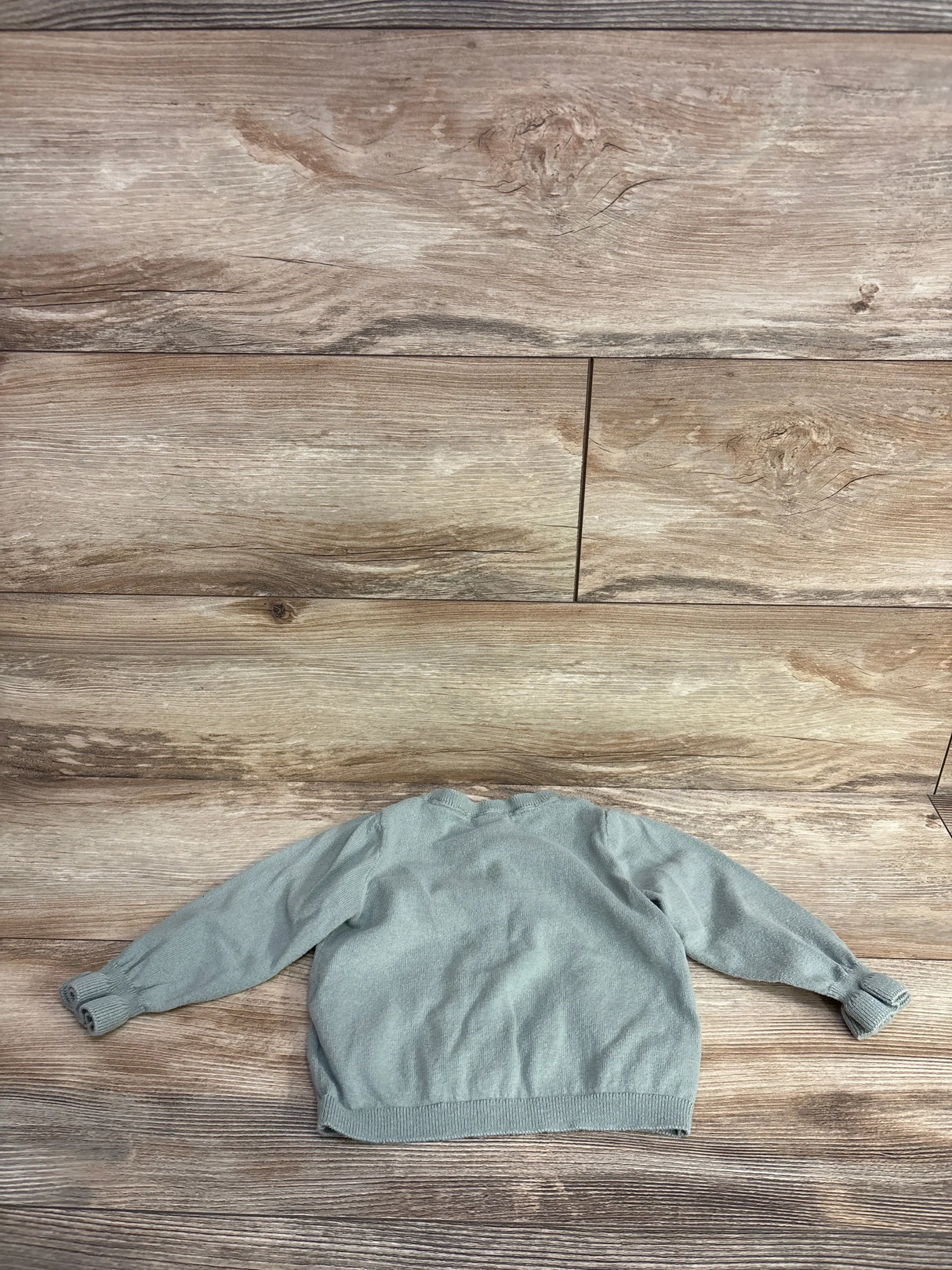 Back view of light green baby button-up cardigan showing knit texture, ribbed cuffs and hem on wood-textured background