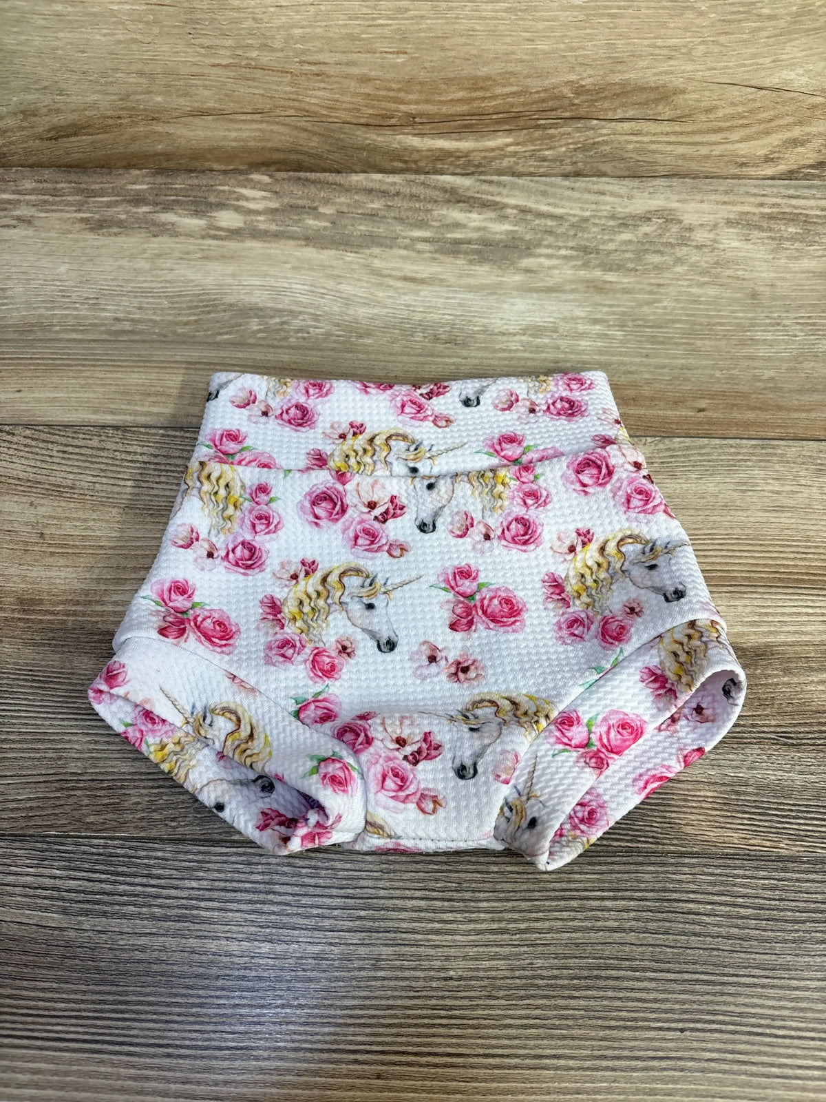 Top-down view of white baby bloomers with unicorn and pink rose print, waffle fabric and elastic waistband on wood