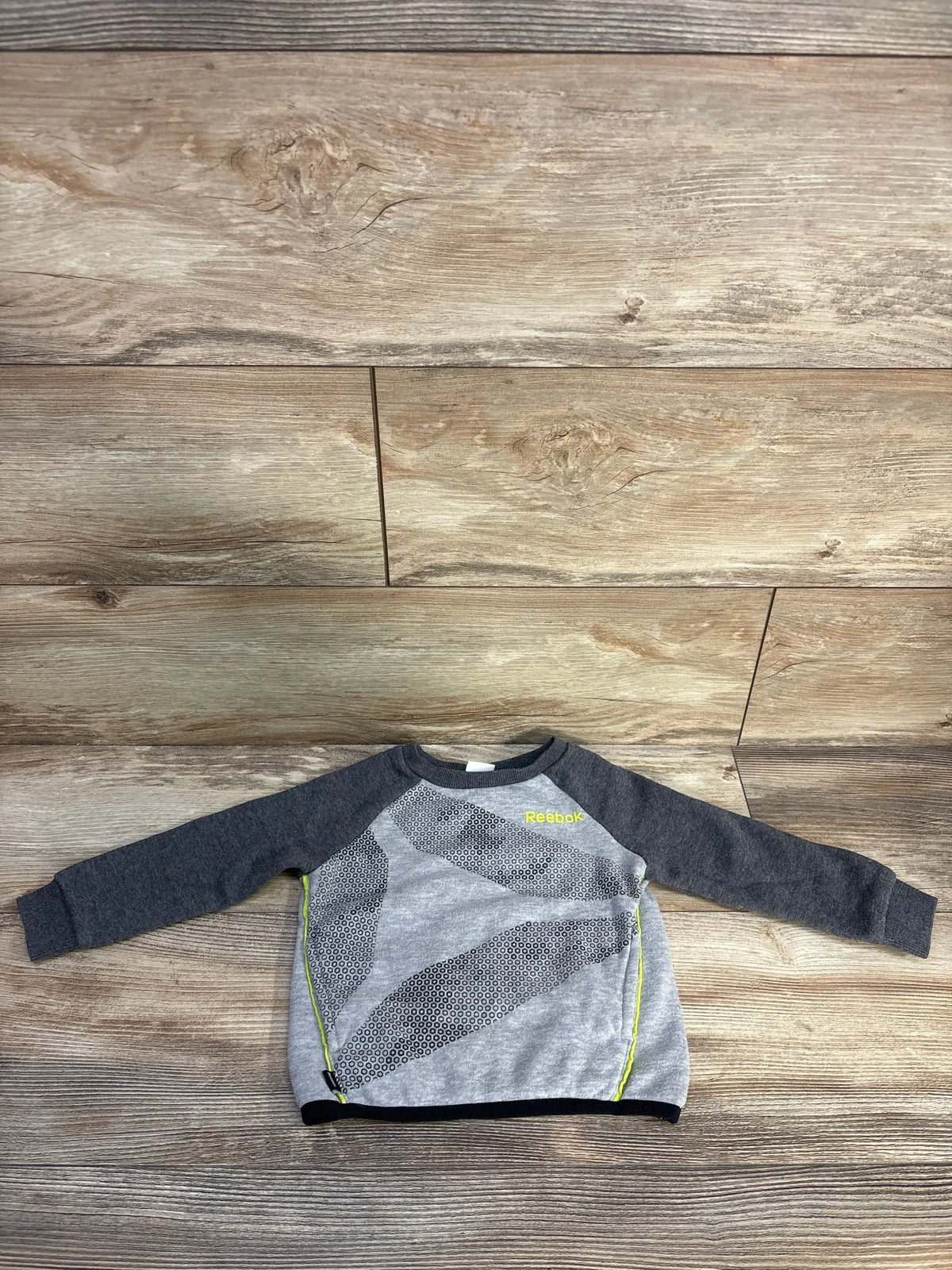 Front view of gray Reebok pullover sweatshirt showcasing dark raglan sleeves, dotted chest graphic, and neon yellow trim on wood flooring