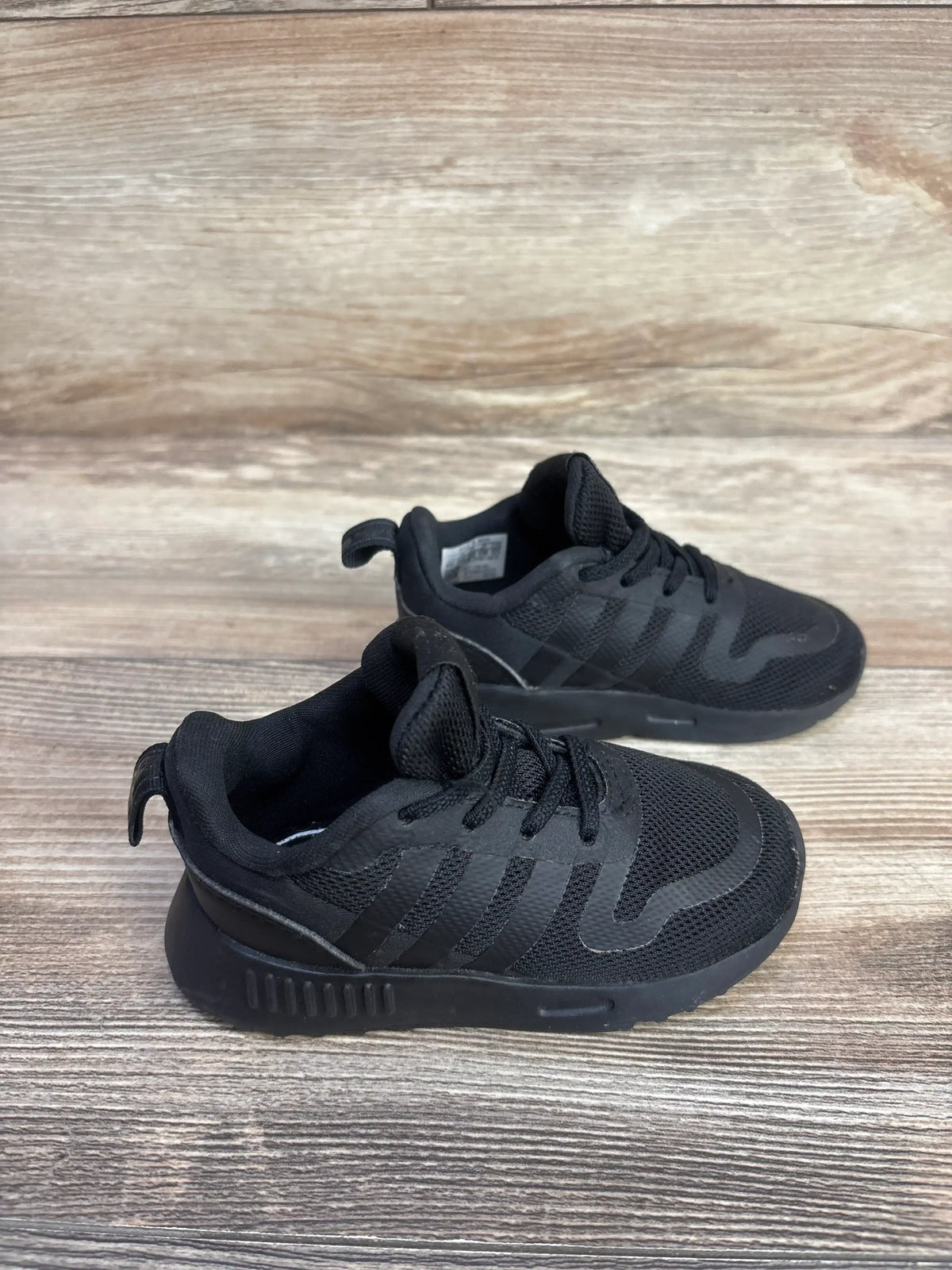 Angled view of Adidas Multix I toddler sneakers in triple black showing mesh upper, three stripes, laces and heel tab on wood background