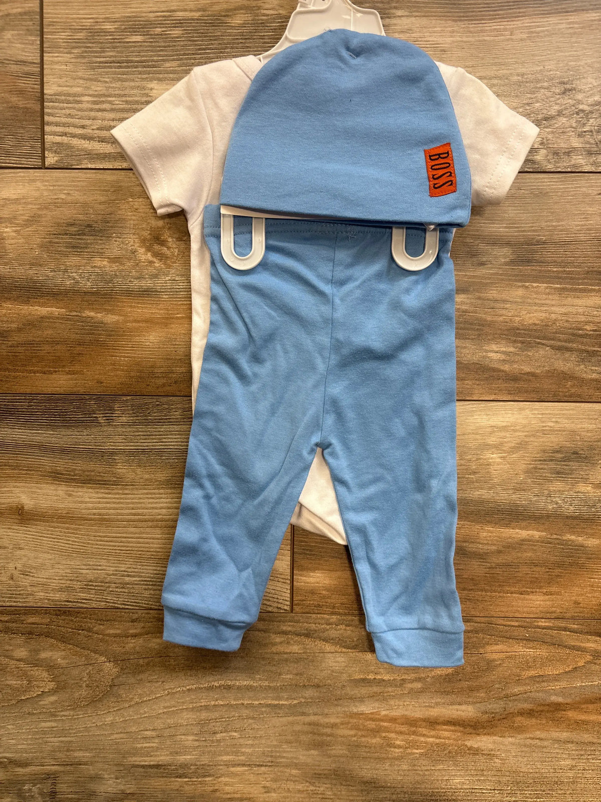 Front view of white short-sleeve bodysuit with light blue pants and matching hat with orange BOSS patch on wood floor