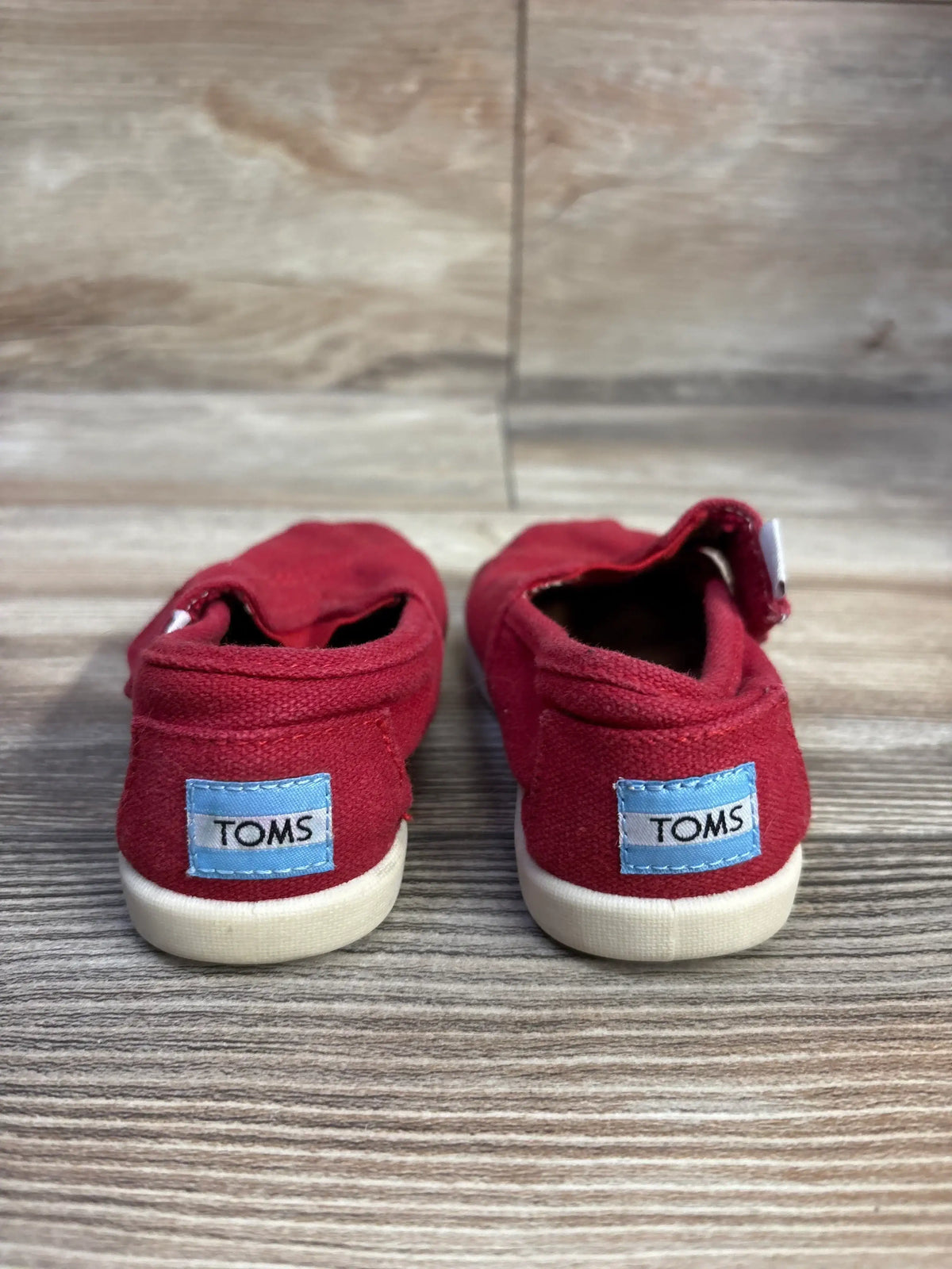 Back view of Toms Tiny Alpargata toddler shoes in red showcasing blue TOMS heel labels and white rubber soles on wood floor