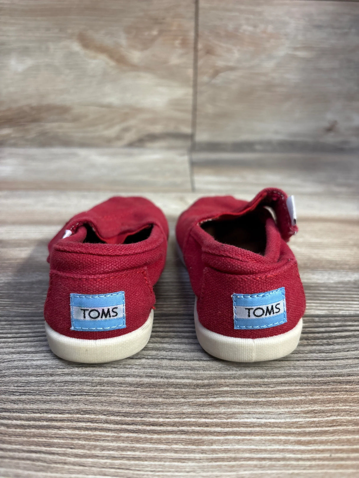 Back view of Toms Tiny Alpargata toddler shoes in red showcasing blue TOMS heel labels and white rubber soles on wood floor