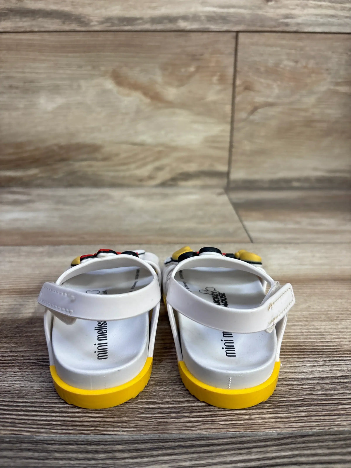 Rear view of Mini Melissa x Disney Minnie & Mickey toddler sandals showing yellow soles and Velcro back straps on wood floor