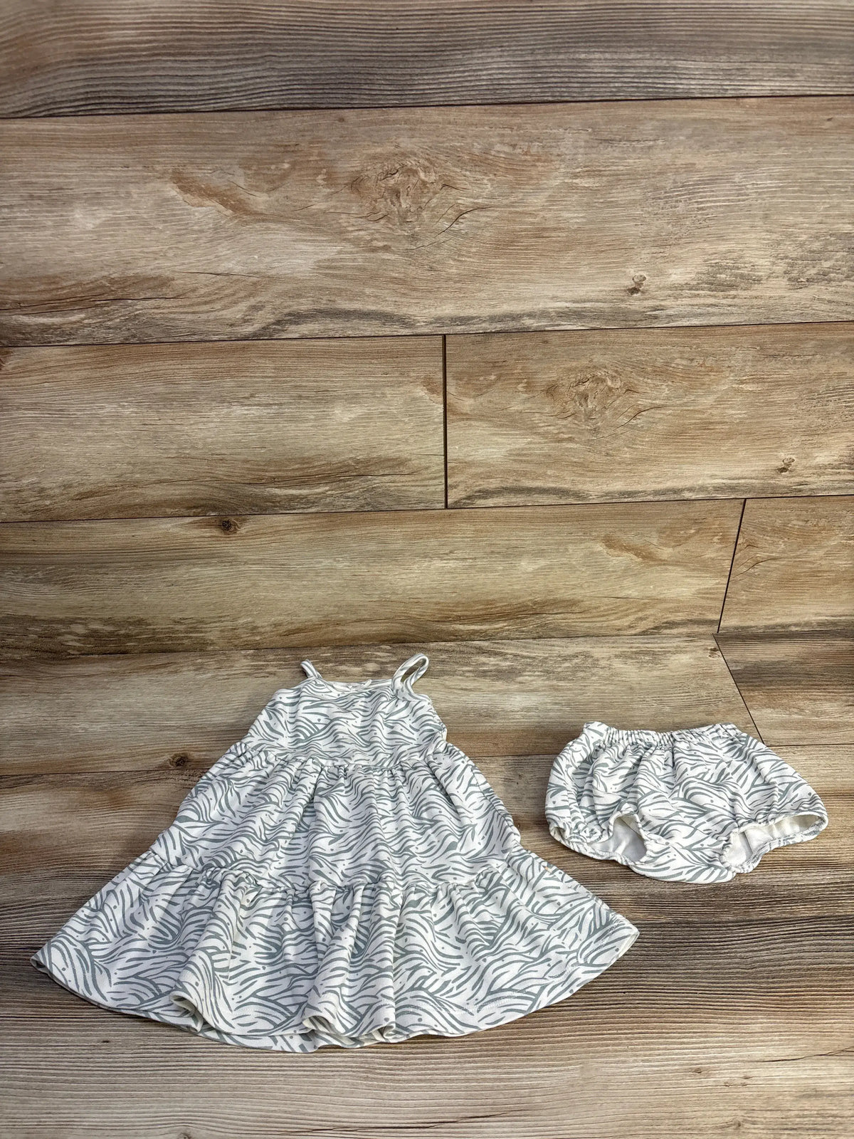 Top-down view of Makemake Organics sleeveless dress and bloomers set showcasing white fabric with gray zebra pattern on a wood floor