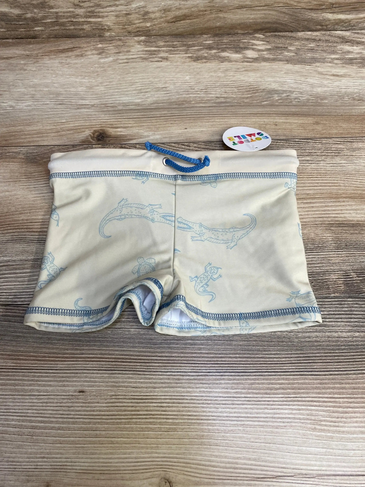 Flat lay of beige children’s swim shorts showcasing light-blue lizard print, blue drawstring and contrast stitching on a wood surface