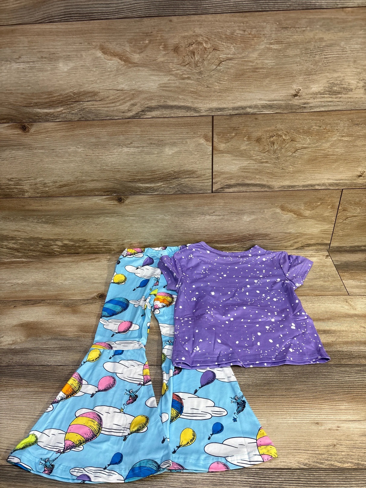 Flat lay of Dr Seuss two-piece toddler set with purple splatter tee and blue hot air balloon bell-bottoms on wood background
