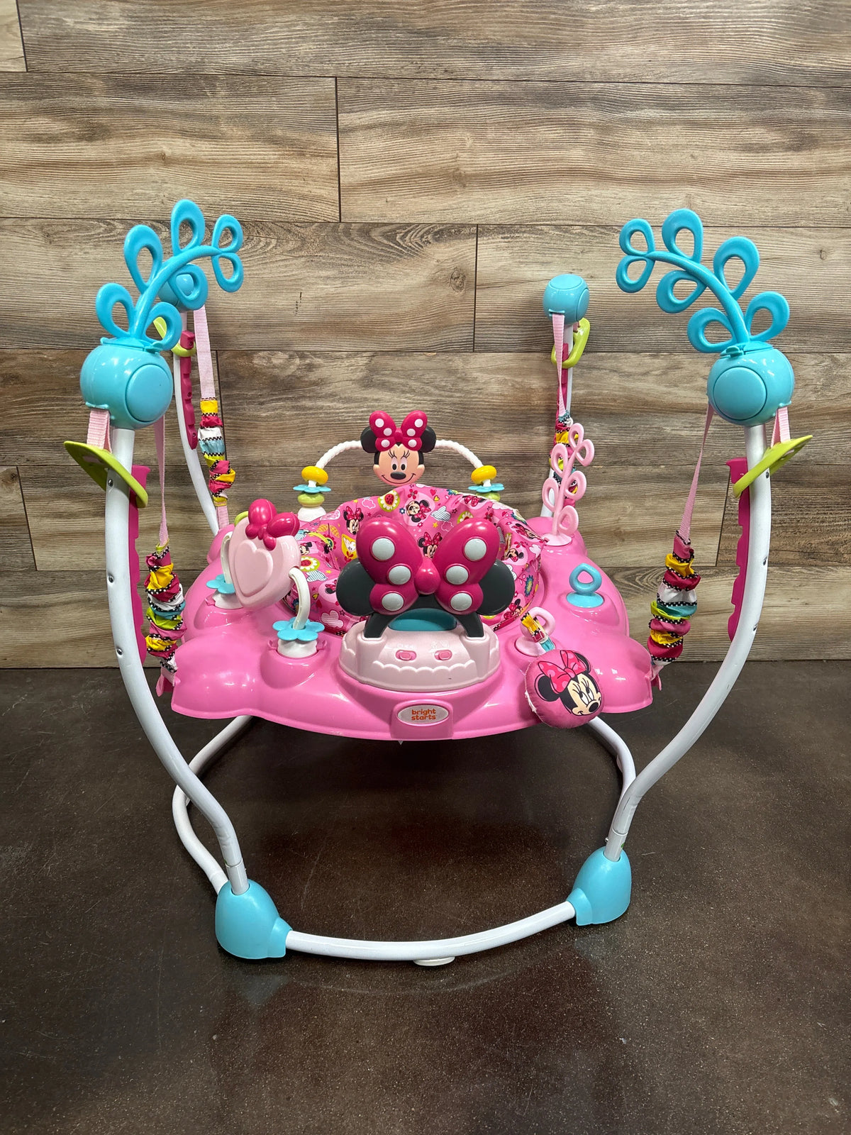 Front view of Disney Baby Minnie Mouse PeekABoo Activity Jumper with pink seat and hanging toys on wood flooring