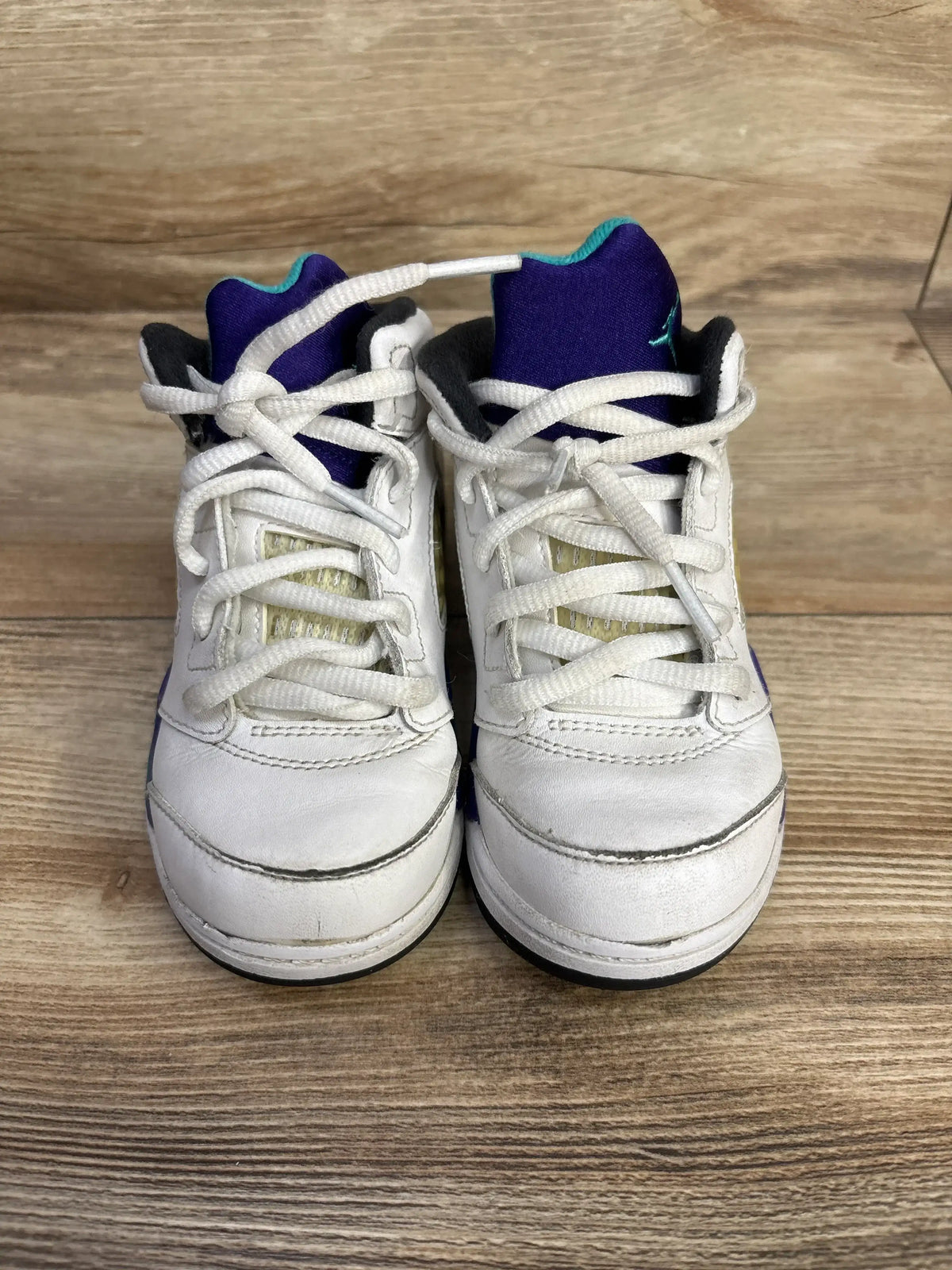 Front of Air Jordan 5 Retro TD Grape sneakers with white uppers, purple tongues, teal Jumpman and white laces on wood floor