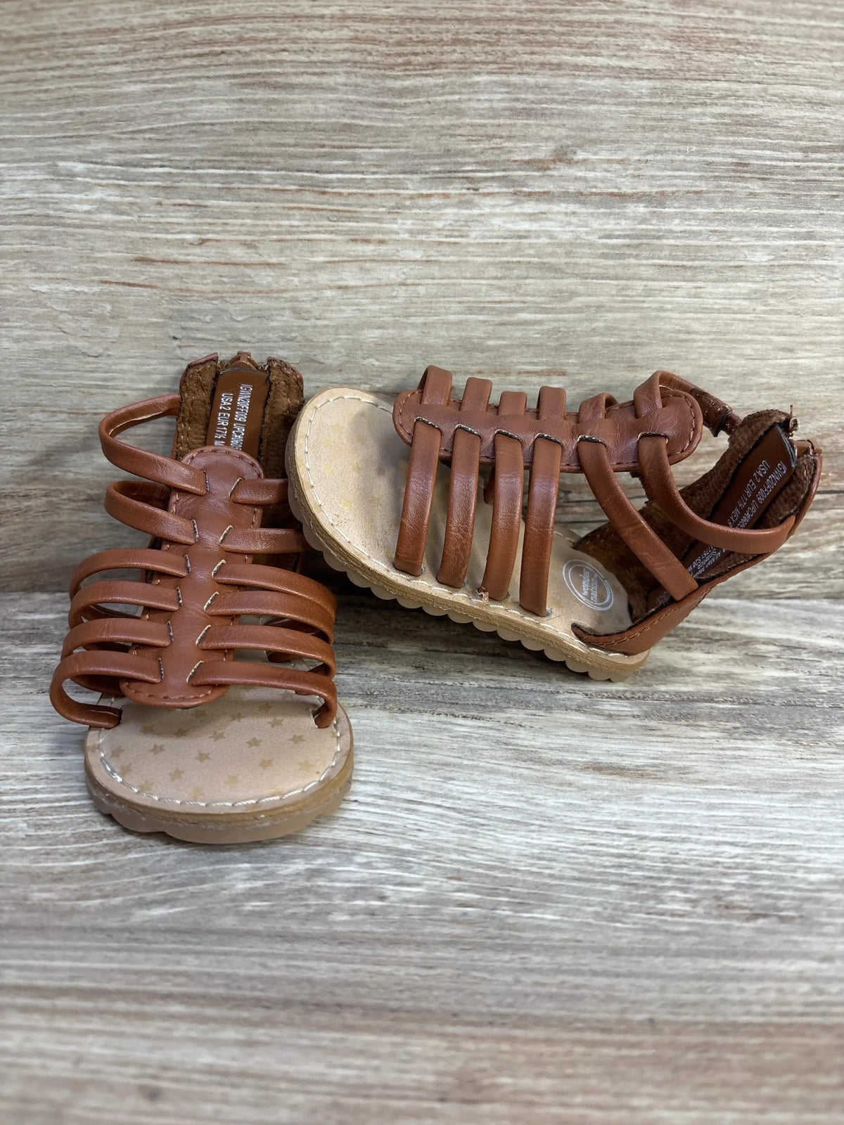 Front and side view of brown toddler gladiator sandals showcasing multiple woven straps and scalloped rubber sole on light wood background