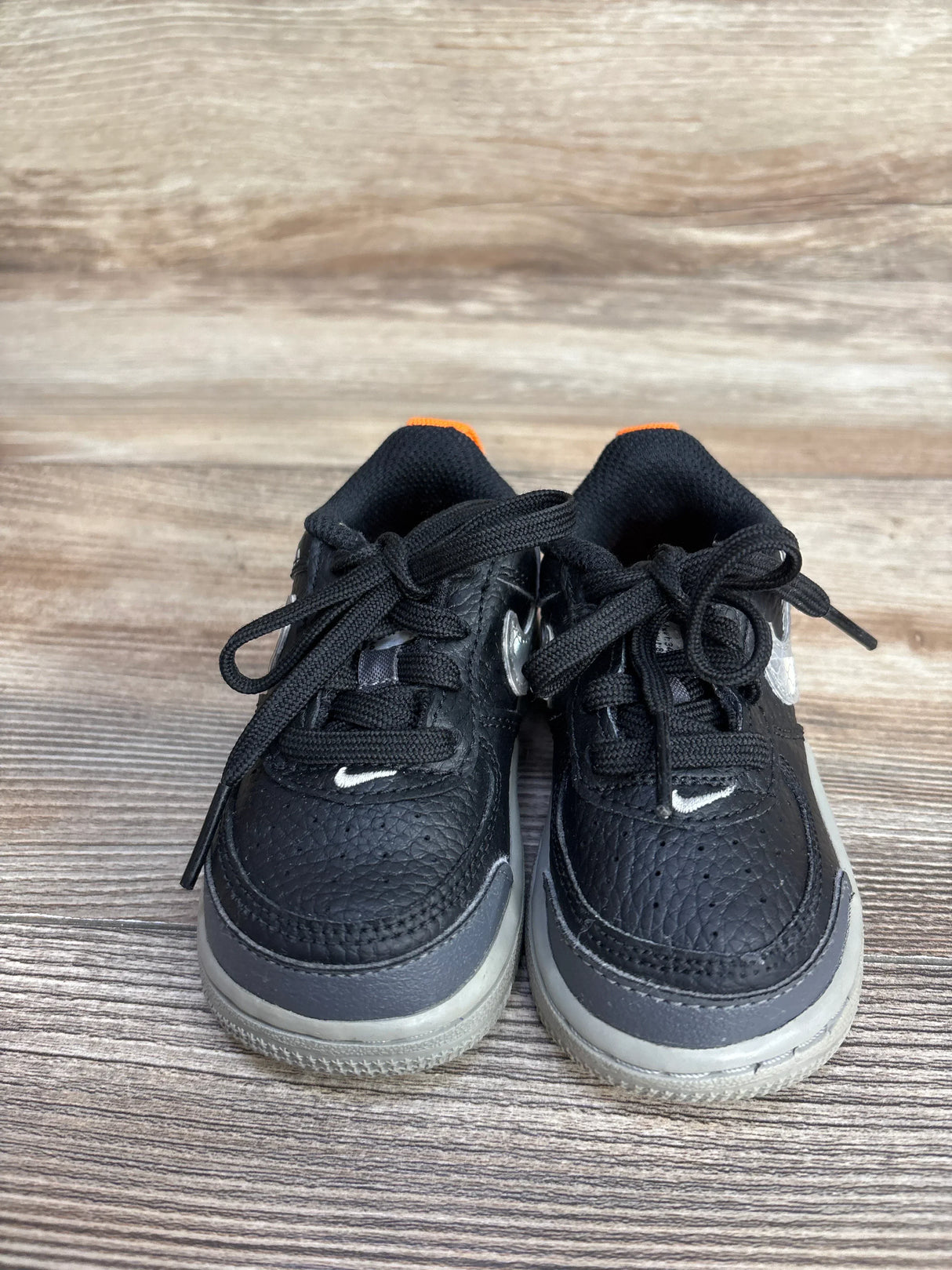 Front view of Nike Air Force 1 toddler sneakers showing black leather, gray soles, white Swoosh and orange heel lining on wood floor