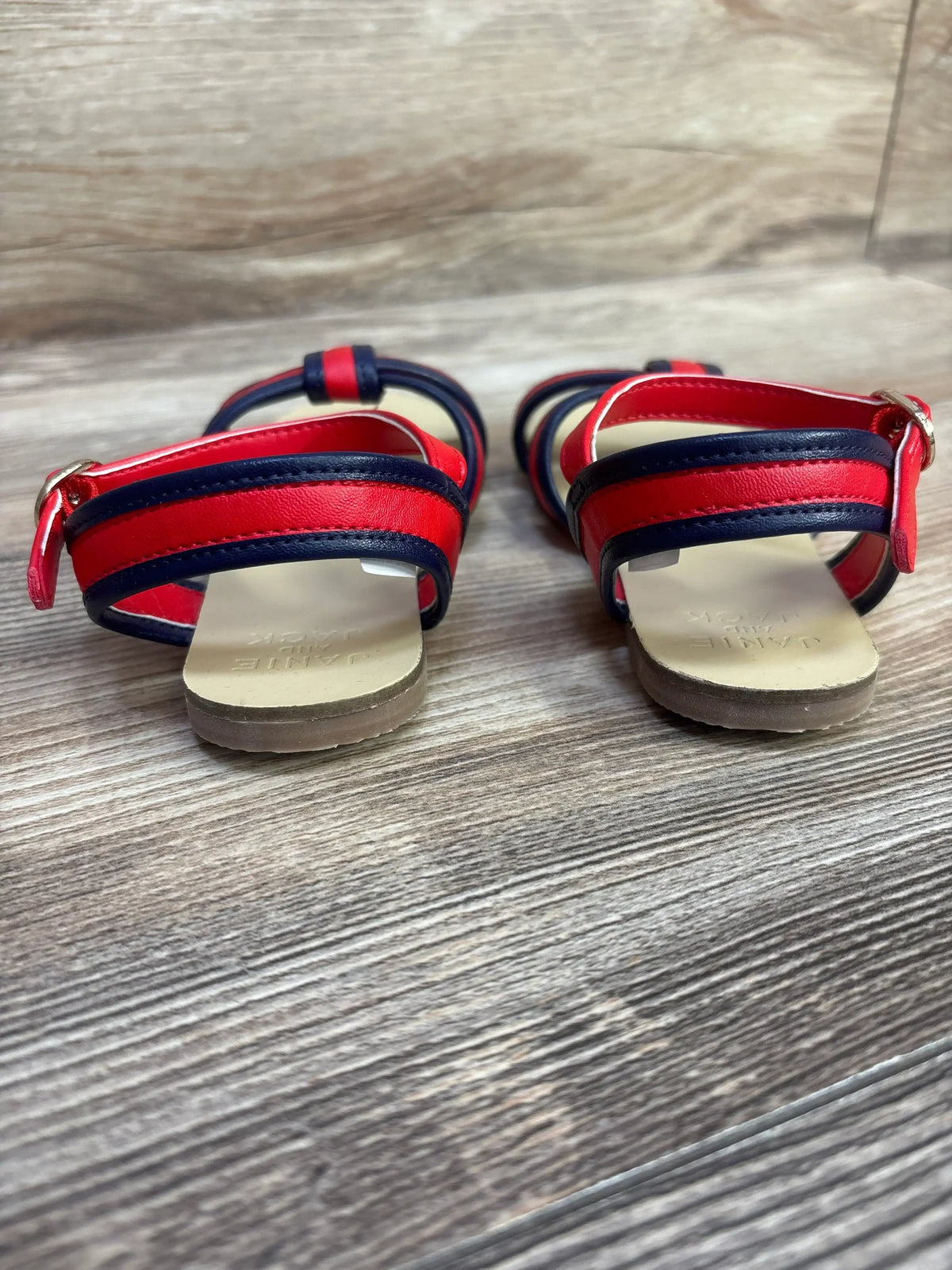 Rear view of red and navy striped cross-strap toddler sandals showing ankle buckle and beige insole on wood floor