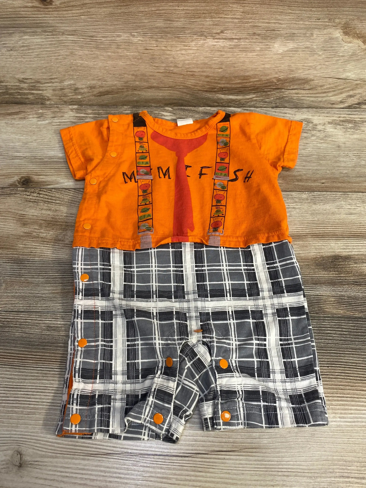 Flat lay of orange Mimi Fish baby romper showing fish-tie print and suspenders, gray plaid bottom with orange snap closures on wood background