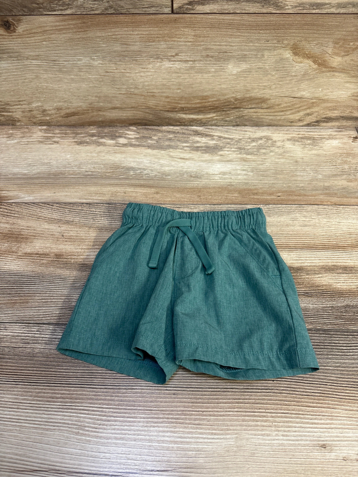 Flat lay of green Tommy Bahama baby board shorts with elastic drawstring waist and side pockets on wood background size 12M