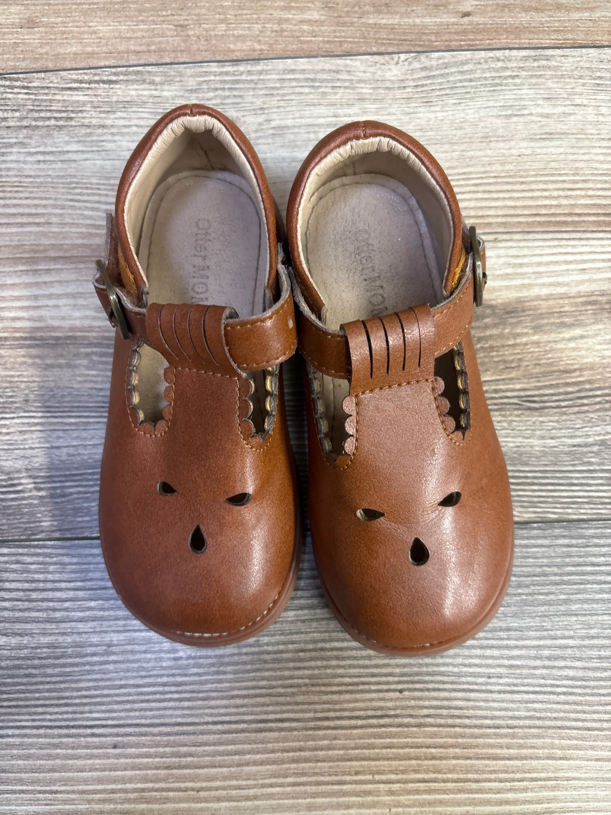 Top view of Otter Momo brown leather T-strap Mary Jane shoes with scalloped strap, teardrop cutouts, buckle on light wood background