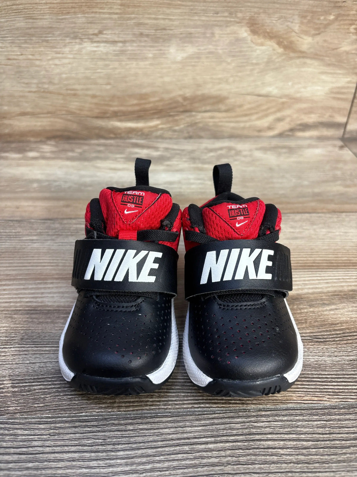 Front view of Nike Team Hustle D8 Black University Red sneakers, size 5C, with Nike straps and white midsoles on wood