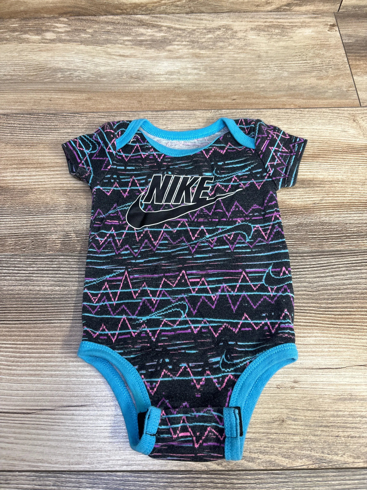 Front view of Nike black baby bodysuit, size 0-6 months, with colorful zigzag pattern and blue trim on a wood floor